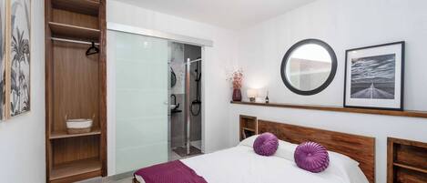 A bedroom with a bed, a wooden wardrobe, a glass shower enclosure, and a round mirror on the wall.