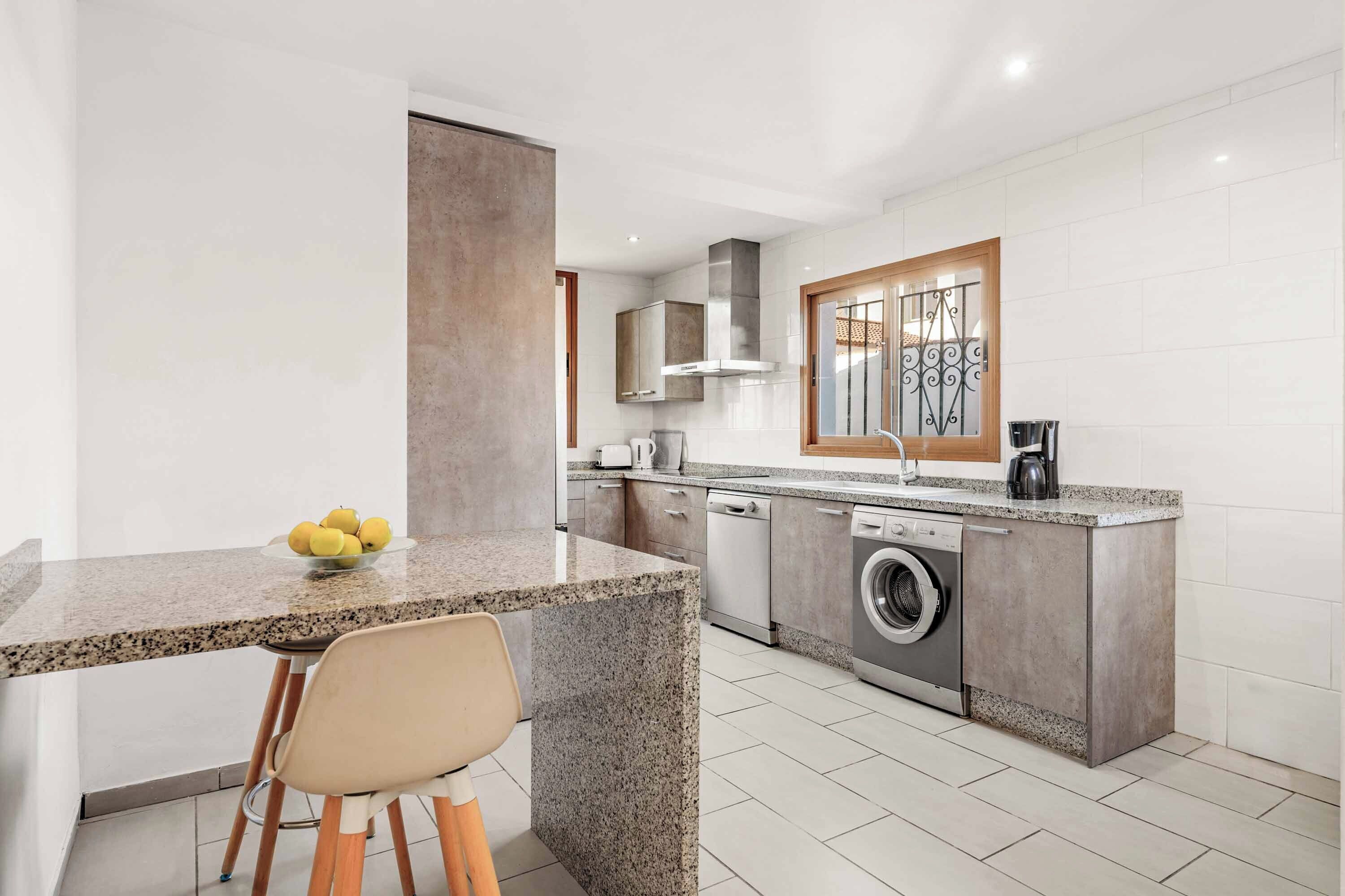 A modern kitchen with granite countertops, a built-in washing machine, and a window with decorative iron bars.