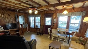 Ein gemütliches Wohnzimmer mit einer Couch, einem Couchtisch und einem Fernseher. Es gibt Fenster mit Vorhängen, einen Ventilator und einen Esstisch mit Stühlen.