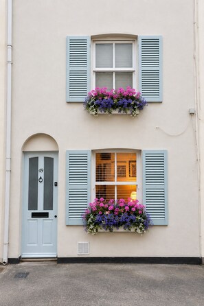 Um edifício com porta azul clara marcada com o número 49, duas janelas com persianas azuis e caixas de flores com flores roxas e rosa.