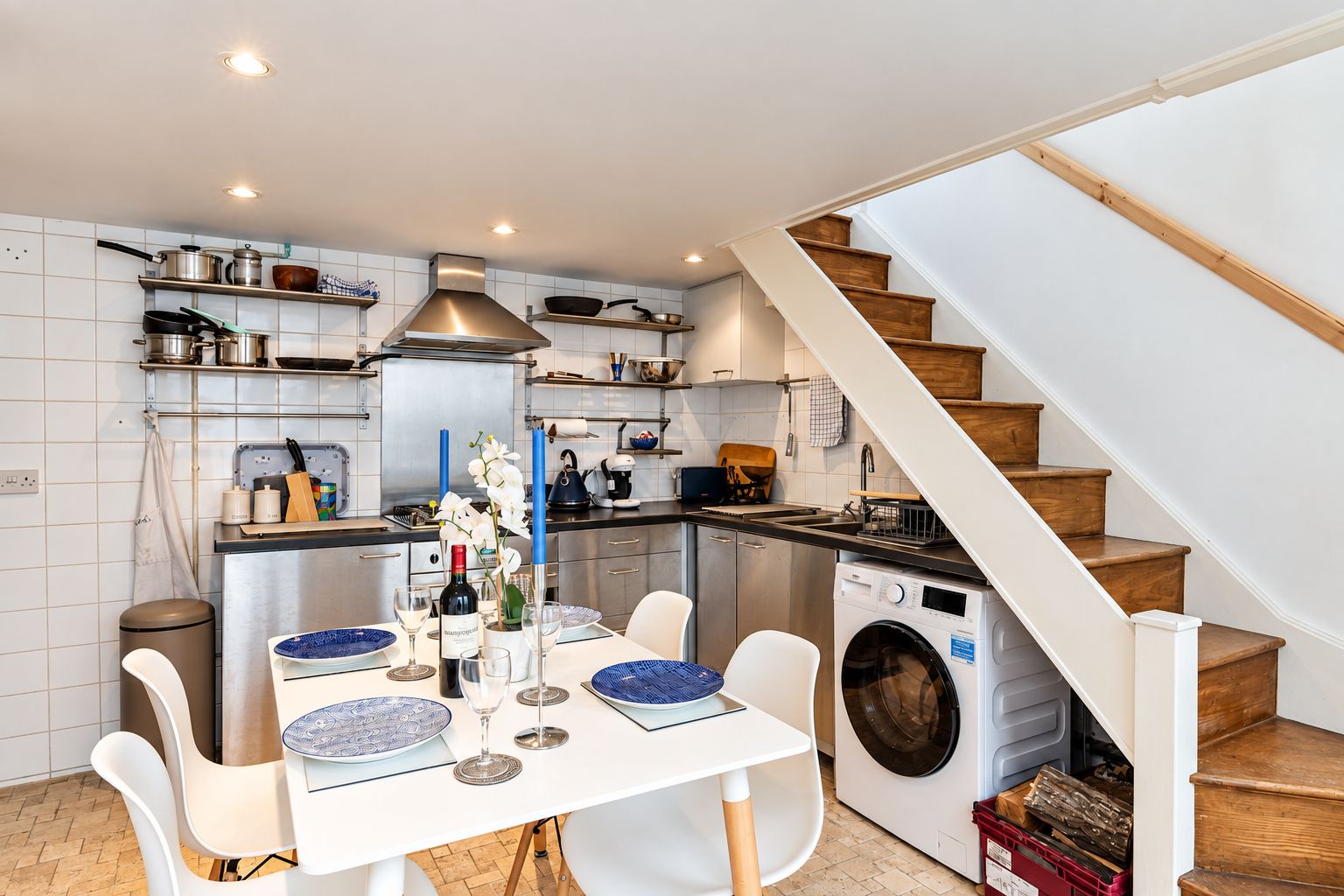 A compact kitchen with a built-in washing machine, a dining area with a table set for four, and a wooden staircase.
