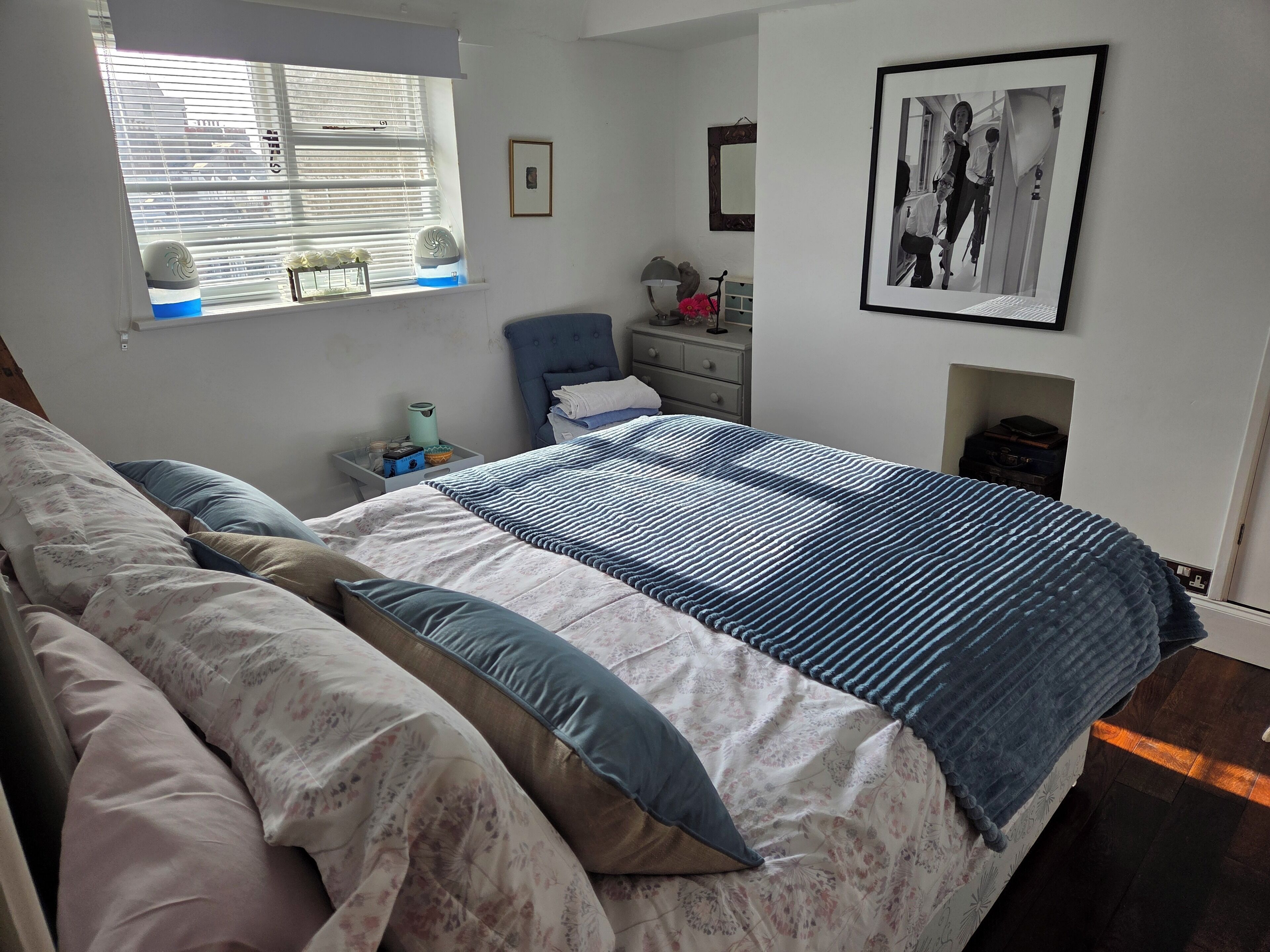 A bedroom with a bed, pillows, a bedside table, a lamp, a framed picture, and a window with blinds.
