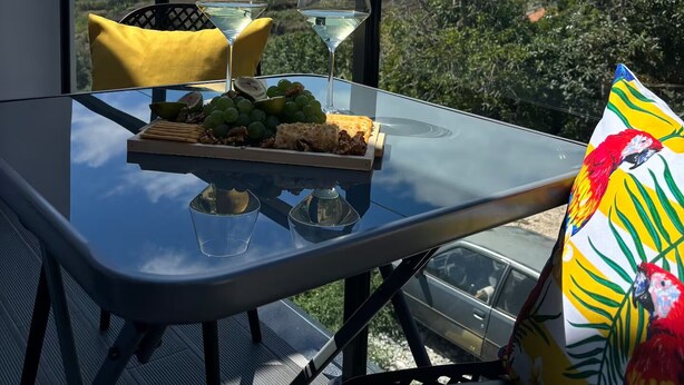 A balcony with a glass table set for two, featuring a bottle of wine, glasses, a plate of grapes, and a small dish of nuts. - Golden Moon, Ponta do Sol - Canhas (Funchal)