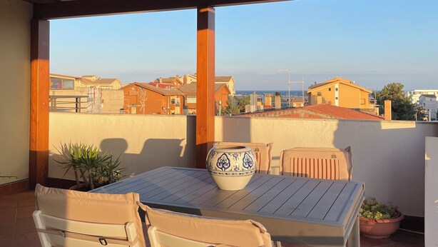 A covered outdoor patio with a table and chairs, a potted plant, and a view of the cityscape. - Delux-The Terrace Over The Marine Park (Quartu Sant'Elena)