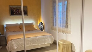 A bedroom with a bed, a bedside table, a mirror, and a window with lace curtains. - Delux-The Terrace Over The Marine Park (Quartu Sant'Elena)
