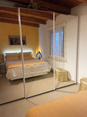A bedroom with a bed, a bedside table, a mirror, and a window with lace curtains. - Delux-The Terrace Over The Marine Park (Quartu Sant'Elena)