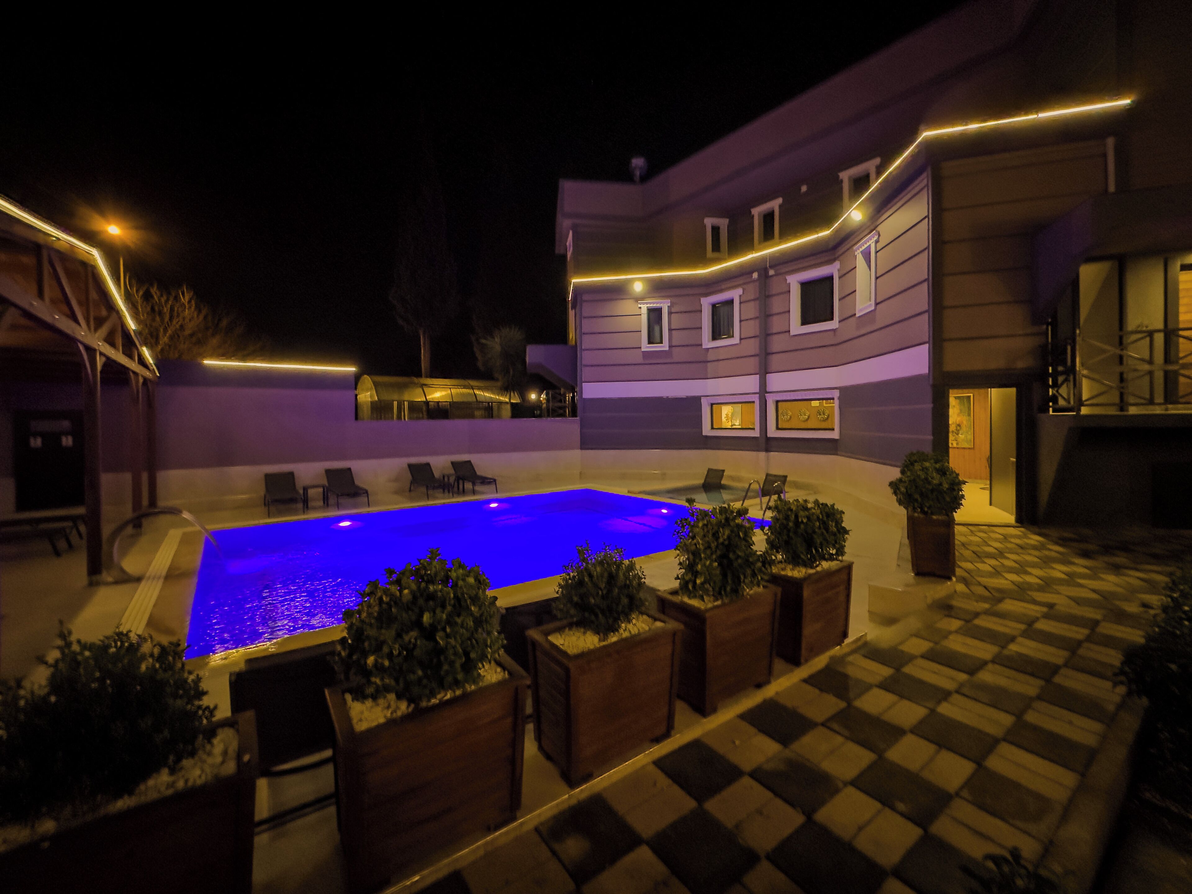 A pool area with lounge chairs and potted plants, surrounded by a building with a checkered floor and illuminated railings.