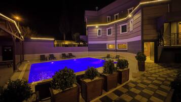 A pool area with lounge chairs and potted plants, surrounded by a building with a checkered floor and illuminated railings.