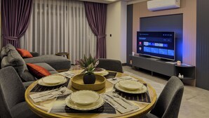 A modern living room with a dining area, featuring a flat-screen TV, a sofa, and a dining table set with plates and cutlery. - Belek 07 Luxury Suites Only For Family (Serik)