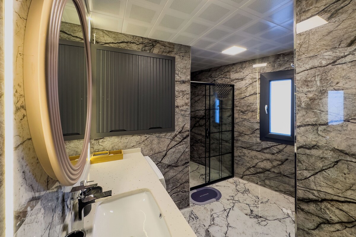 A modern bathroom with a large mirror, marble walls, a bathtub, and a glass shower enclosure.