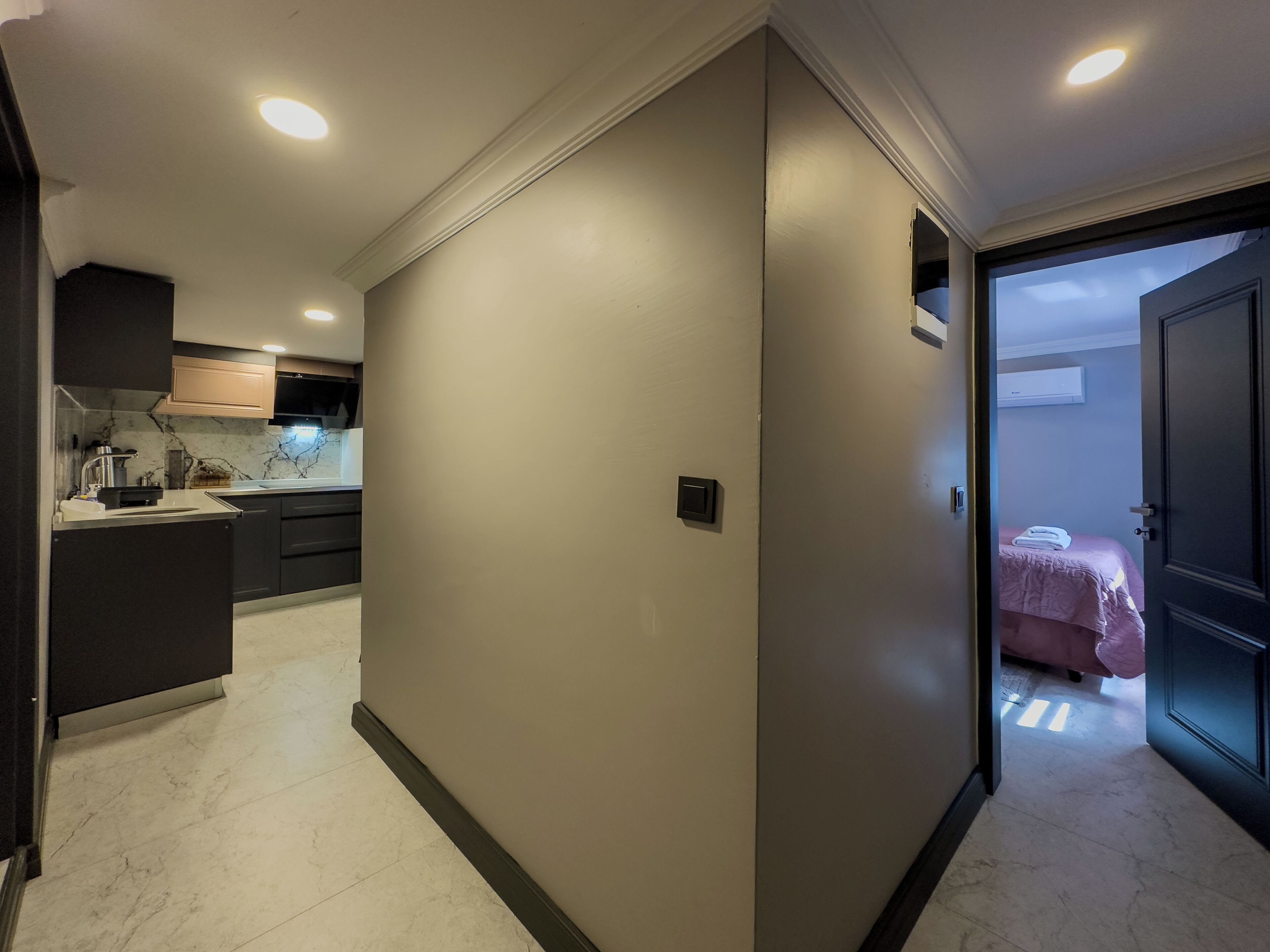 A hallway with a kitchen area on the left, a door leading to a bedroom with a bed and a pink bedspread, and a white air conditioning unit on the wall.