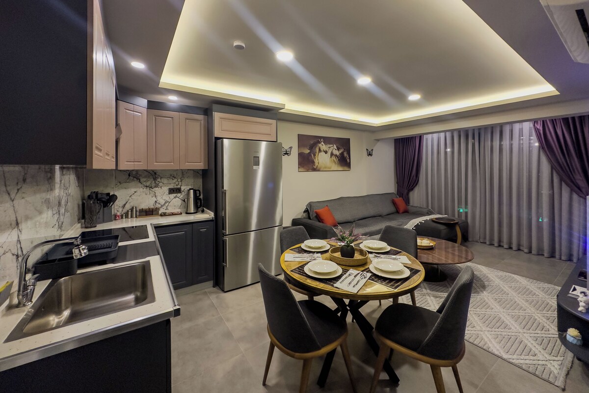 A modern kitchen with a built-in refrigerator, a dining area with a round table and four chairs, and a living area with a sofa and curtains.