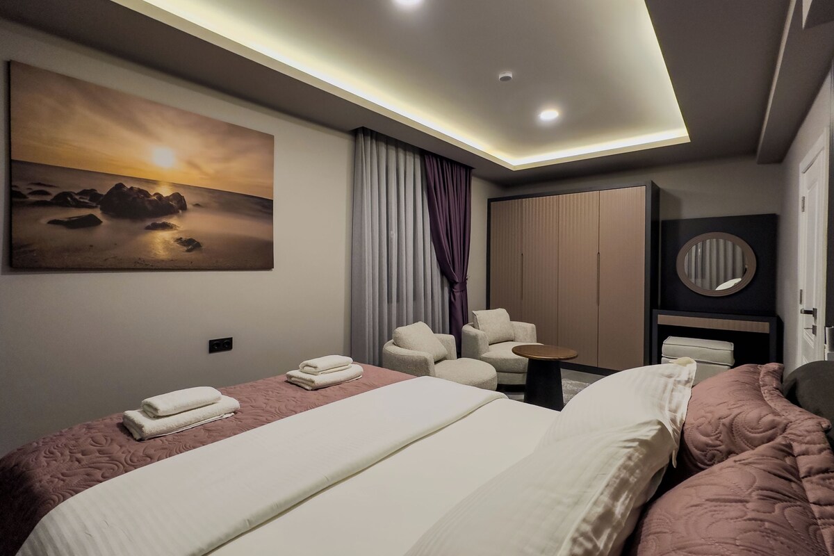 A modern hotel room with a large bed, two armchairs, a small table, a wardrobe, and a painting of a sunset.
