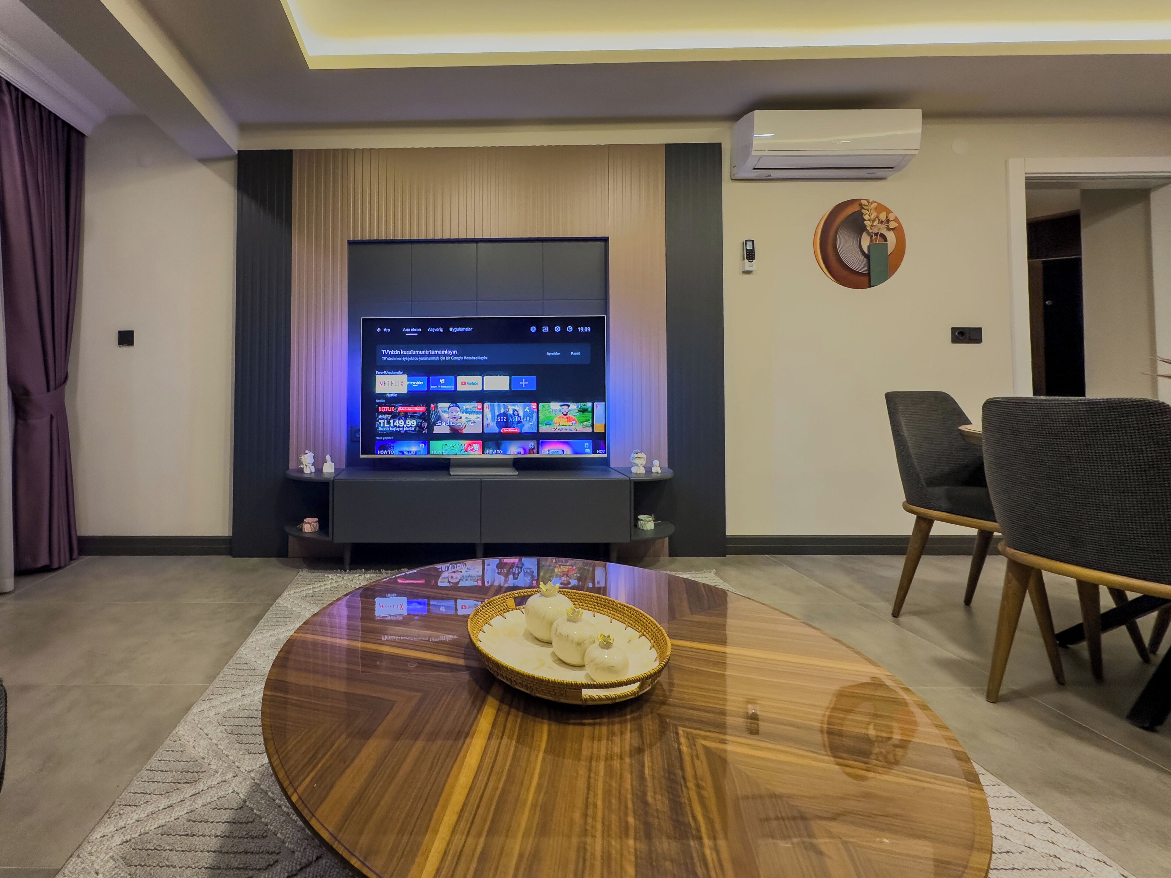 Ein modernes Wohnzimmer mit einem an der Wand befestigten Flachbildfernseher, einem runden Couchtisch mit dekorativer Schale und zwei Sessel.