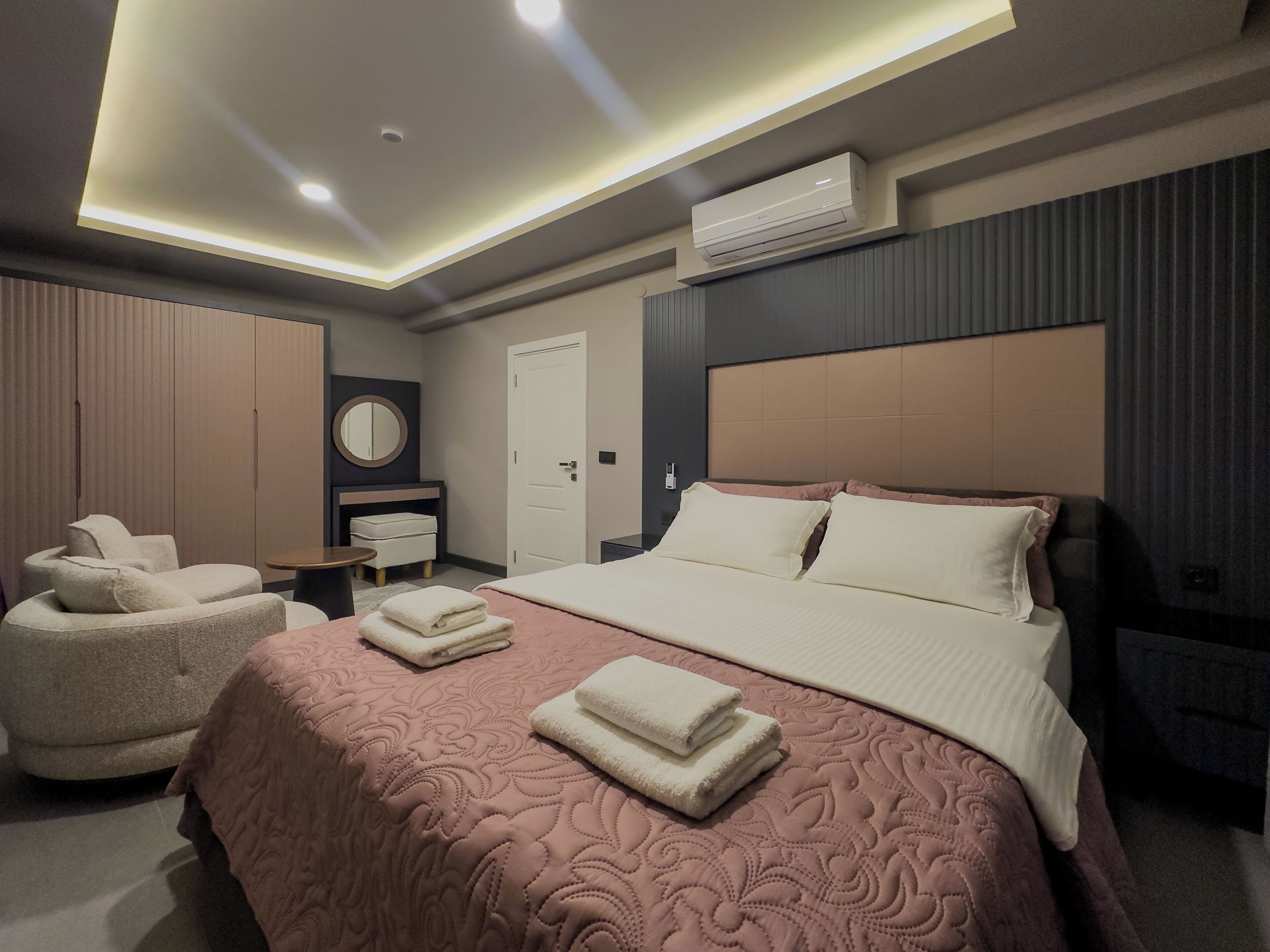 A modern hotel room with a large bed, a seating area with a sofa and armchairs, a vanity with a mirror, and an air conditioning unit on the wall.