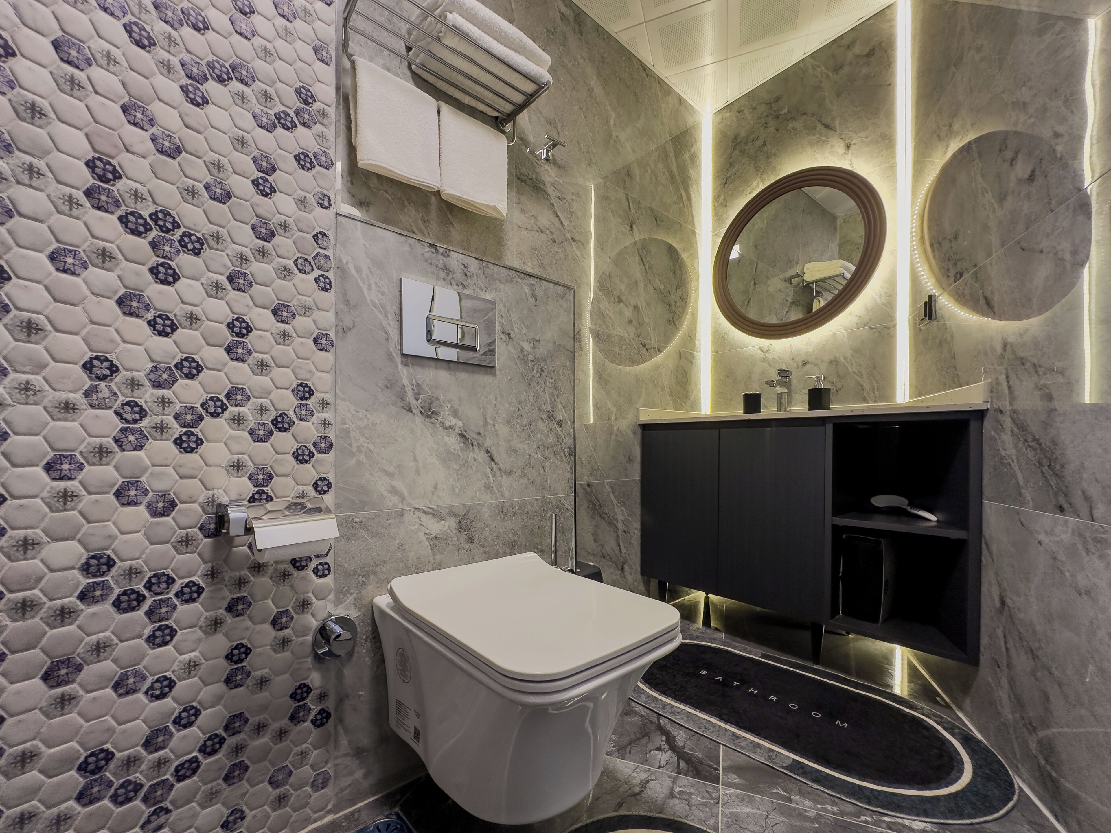 Ein modernes Badezimmer mit Rückwand aus sechseckigen Fliesen, einer weißen Toilette, einem schwarzen Waschtisch mit Spiegel und einer Fußbodenheizmatte mit der Aufschrift 'BADEZIMMER'.
