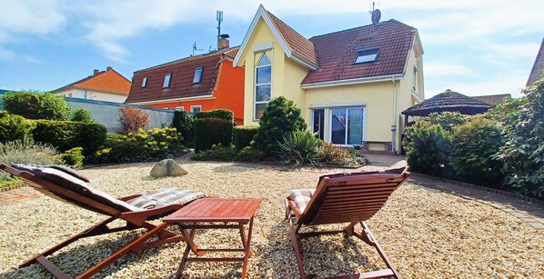Ein Haus mit Kiesterrasse, zwei Holzliegen und einem kleinen Tisch.