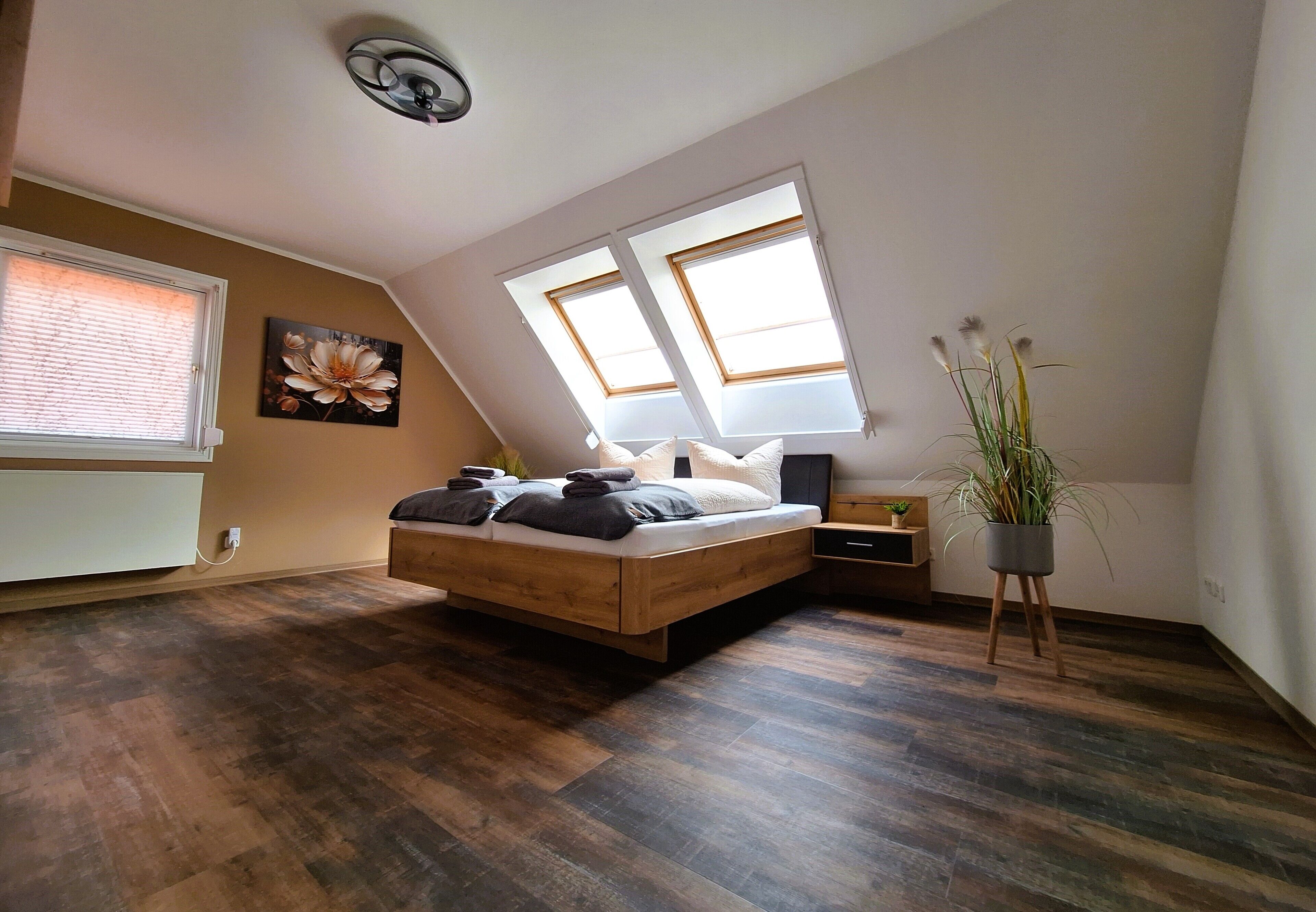 Een slaapkamer met een schuine kap, een bed met kussens, een nachtkastje, een vloerlamp en een raam met jaloezieën.