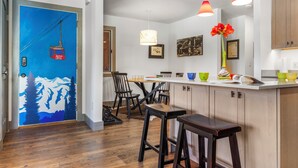 Eine moderne Küche mit Holzschränken, ein Essbereich mit Tisch und Stühlen sowie eine farbenfrohe Tür mit Bergmotiv.