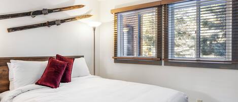 Un dormitorio con cama, ropa de cama blanca, dos almohadas rojas, cabecera de madera, una lámpara de pie y una ventana con persianas.