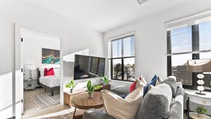 A modern living room with a sofa, a coffee table, and a TV. There are windows with blinds, a door, and a small table with plants.