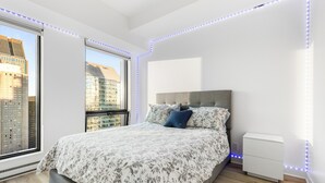 Une chambre moderne avec un grand lit, une table de chevet et une vue sur le paysage urbain, visible à travers une grande fenêtre.