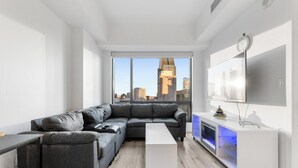Un salon moderne avec un canapé d’angle gris, une table basse blanche, un téléviseur posé sur un meuble blanc et une vue sur la ville à travers de grandes fenêtres.