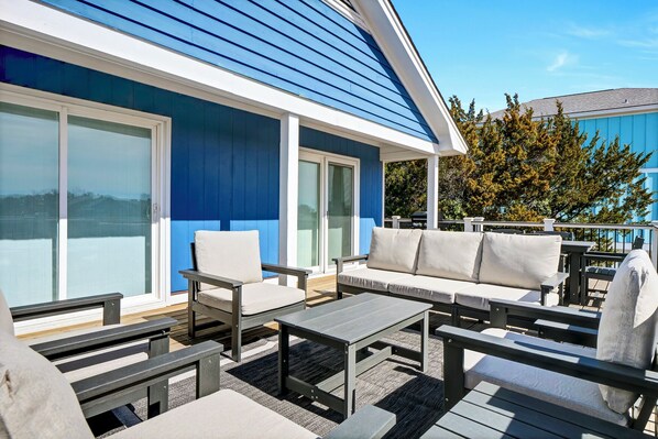 A covered outdoor seating area with a blue building in the background. - The Sea Guld: Beach Access, Expansive Decks, Elevator & Outdoor Shower! (Wrightsville Beach)