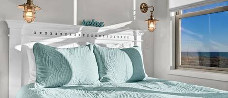 A bedroom with a white headboard, two wall-mounted lights, a bed with a light blue quilt, and a window with a view of the sea.