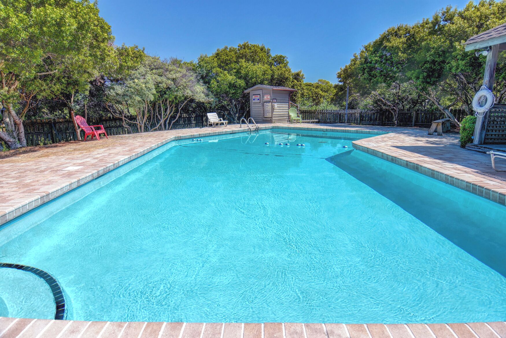 A rectangular swimming pool with a tiled edge, surrounded by a brick patio. There are lounge chairs and a small shed in the background.