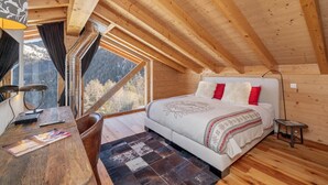 Ein Schlafzimmer mit Holzbalkendecke, einem Bett mit weißer Bettwäsche und roten Kissen, einem Schreibtisch mit Lampe und Blick auf einen Wald durch ein Fenster.