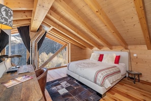 Ein Schlafzimmer mit Holzbalkendecke, einem Bett mit weißer Bettwäsche und roten Kissen, einem Schreibtisch mit Lampe und Blick auf einen Wald durch ein Fenster.