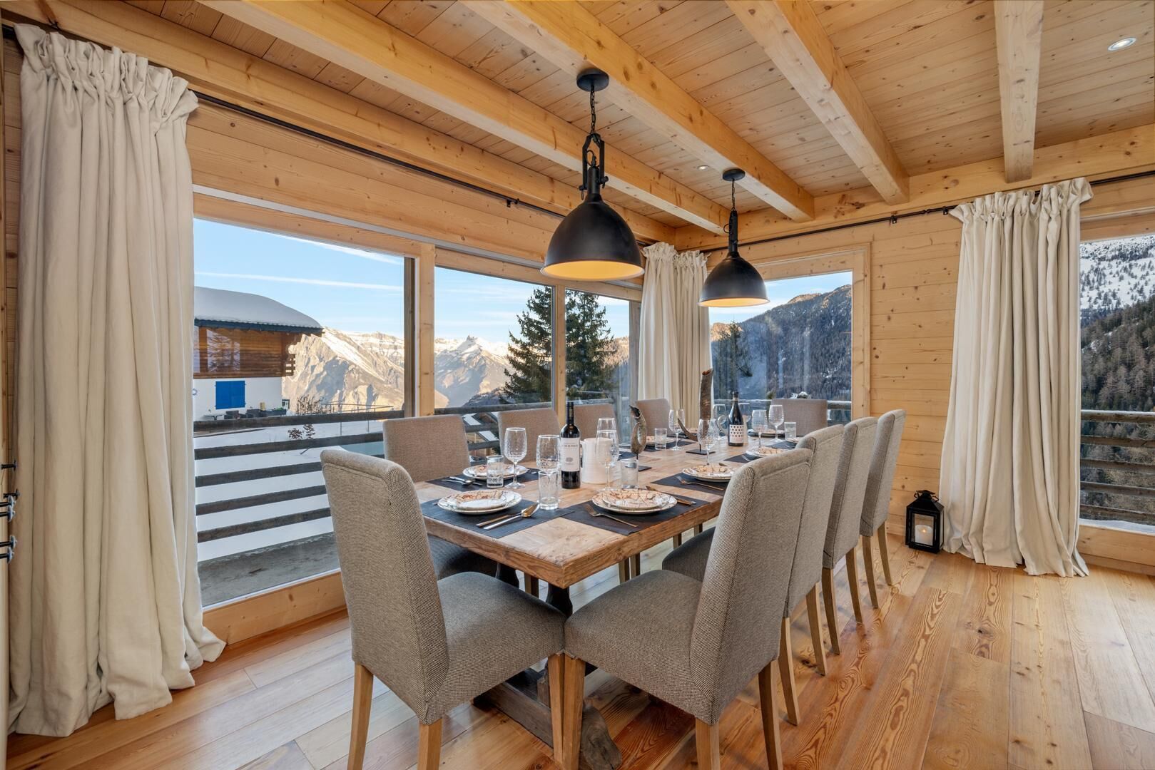 Comedor de madera con una mesa dispuesta para cuatro, ventanas amplias con cortinas y vistas a montañas.