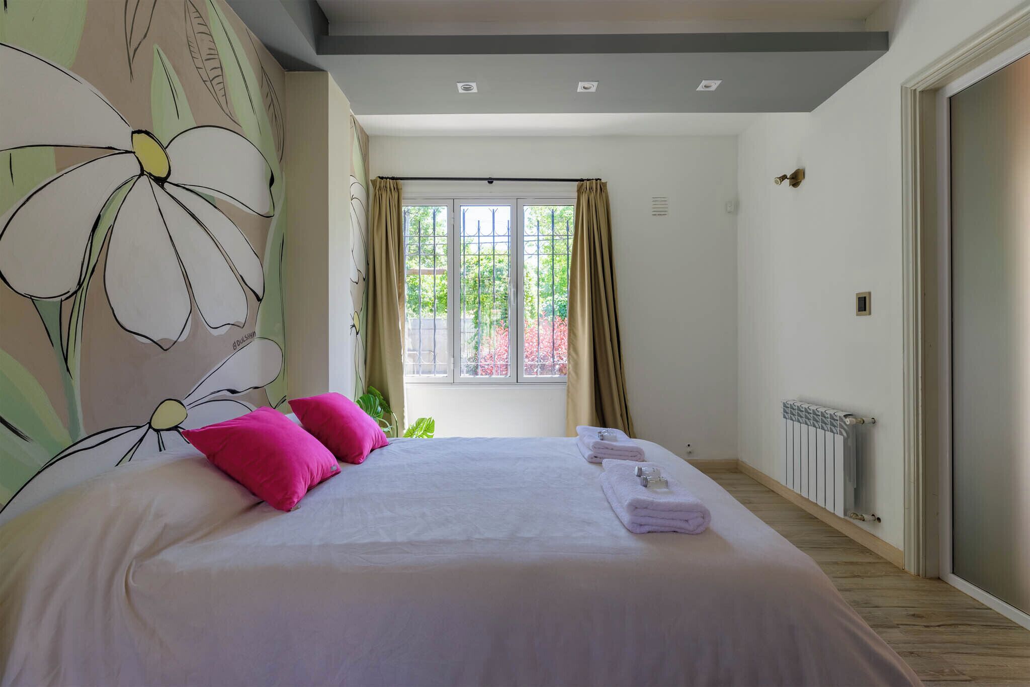 Une chambre à coucher avec un grand lit, deux oreillers roses, une fenêtre avec des rideaux et un radiateur.