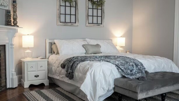 Un dormitorio con cama, dos mesitas de noche, cómoda, chimenea y dos ventanas con adornos.