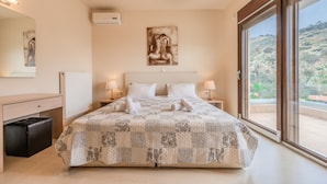 Un dormitorio con cama, dos mesitas de noche, un tocador, cómoda y un espejo, junto a un amplio ventanal con vistas a un paisaje montañoso.