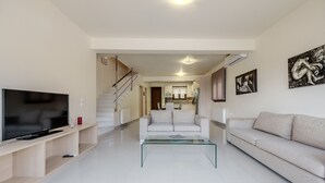Una sala de estar moderna con un televisor de pantalla plana, una mesa de centro de vidrio, un sofá y una escalera que lleva a una cocina.