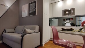 Un salon moderne avec un canapé gris, un fauteuil rose et un coin cuisine doté de meubles blancs et d’un four à micro-ondes.
