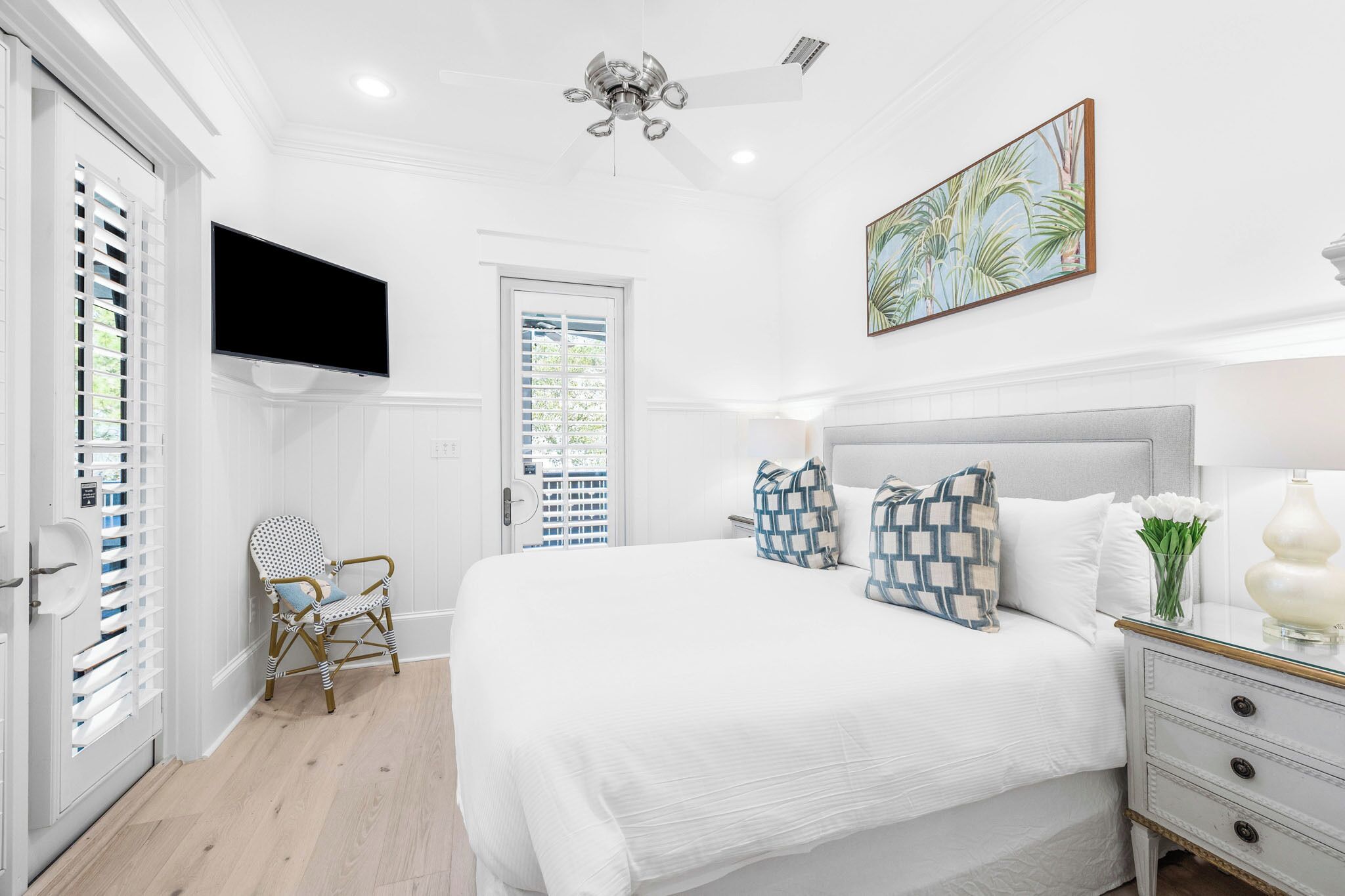A bedroom with a bed, a television mounted on the wall, a ceiling fan, and a window with blinds.