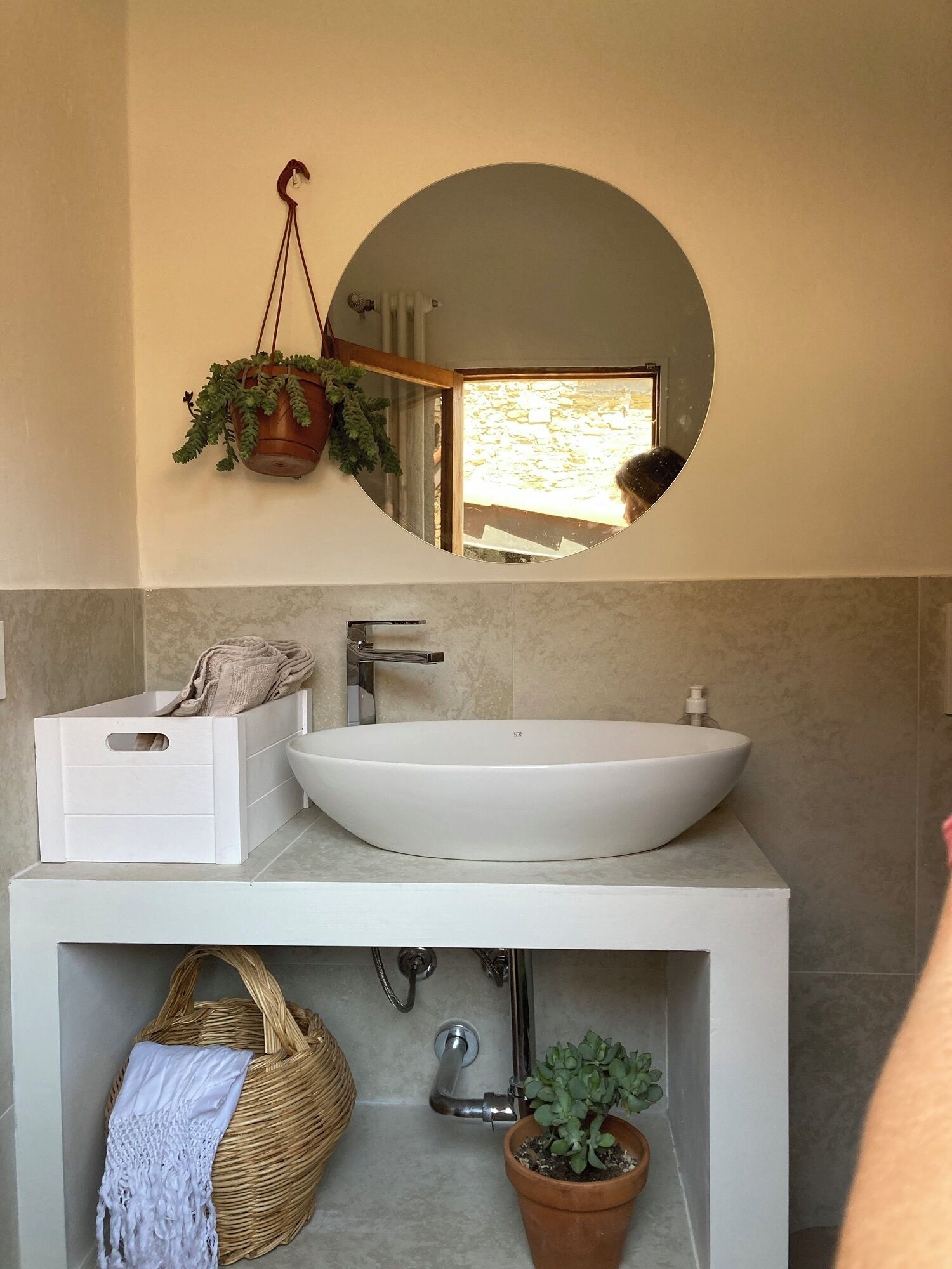 Een badkamer met een witte wastafel, een spiegel, een hangplant, een rieten mand en een bloempot.