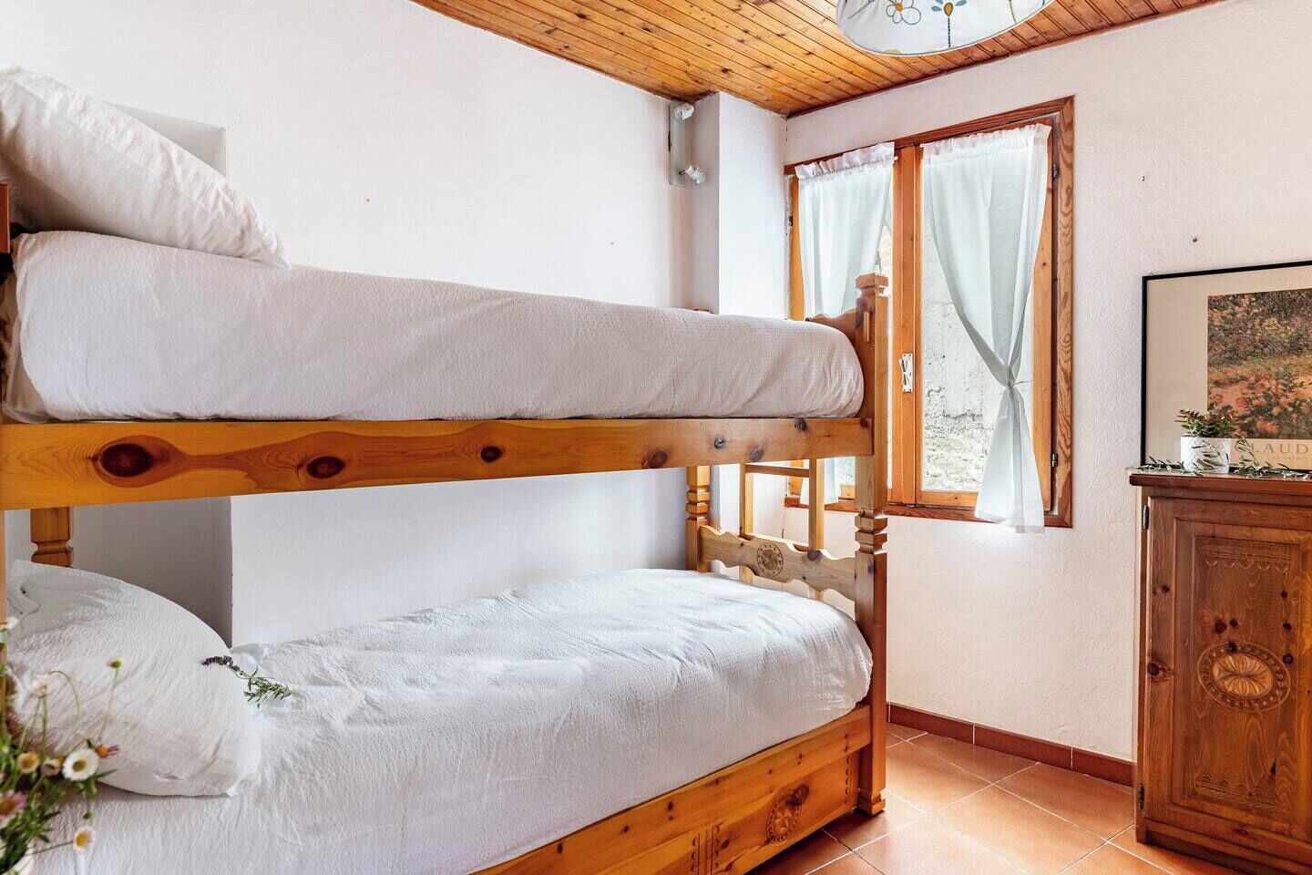 Un lit superposé avec une literie blanche, dans une pièce dotée de poutres en bois et d’une fenêtre avec des rideaux.