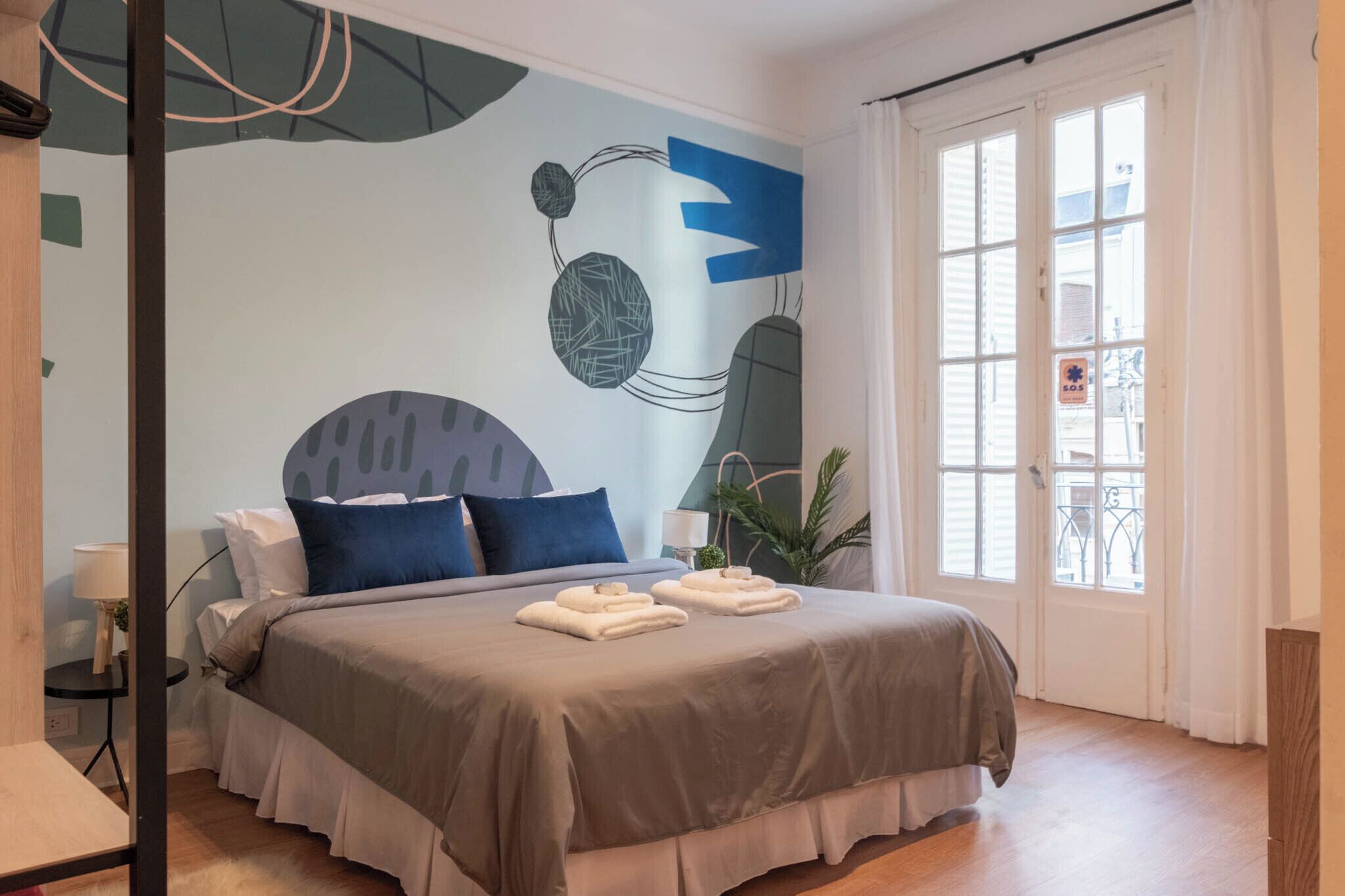 Un dormitorio con cama, almohadas, una mesita de noche, lámpara y ventana con cortinas.