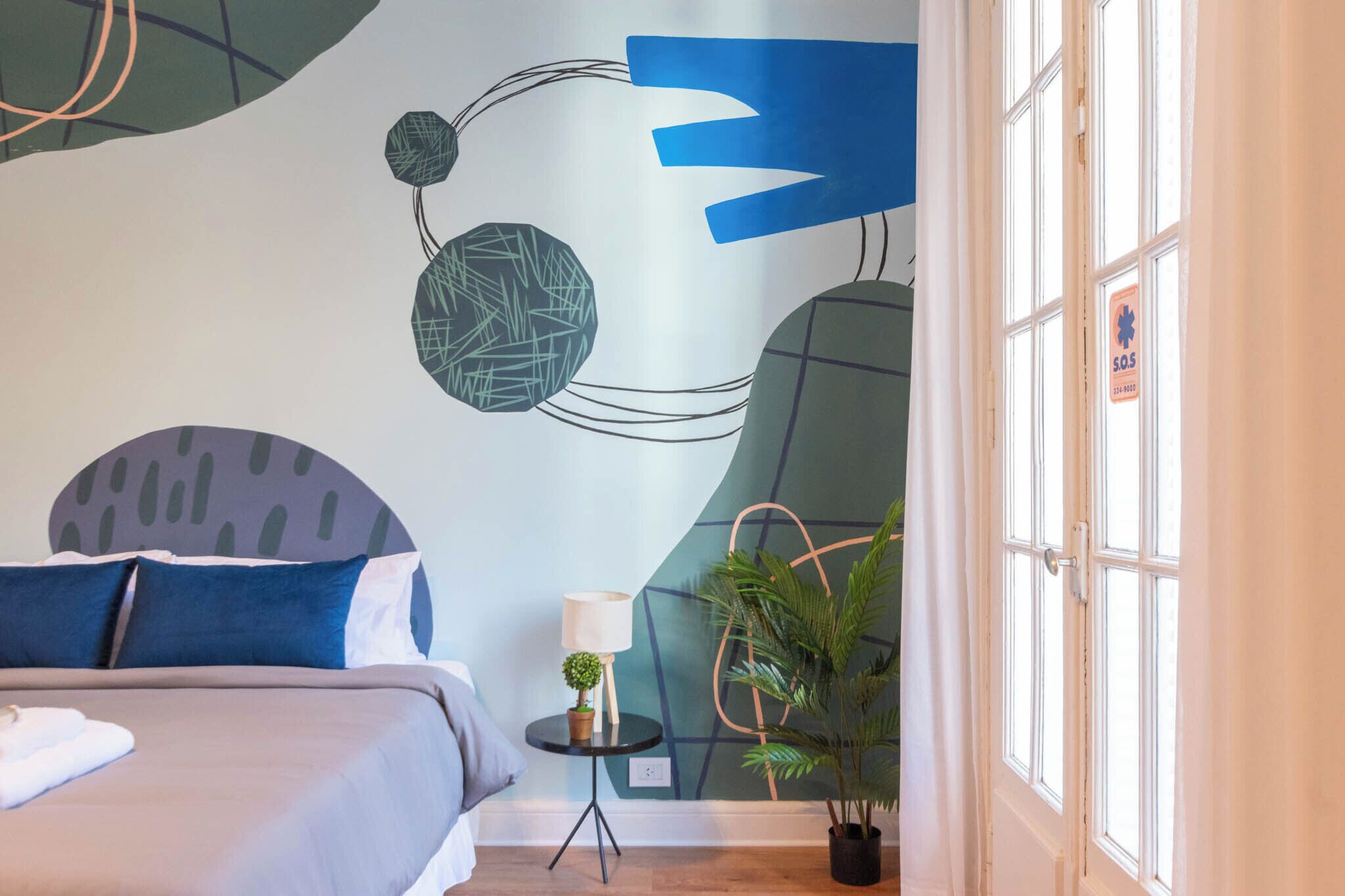 A modern hotel room with a bed, a small table with a lamp, and a potted plant.