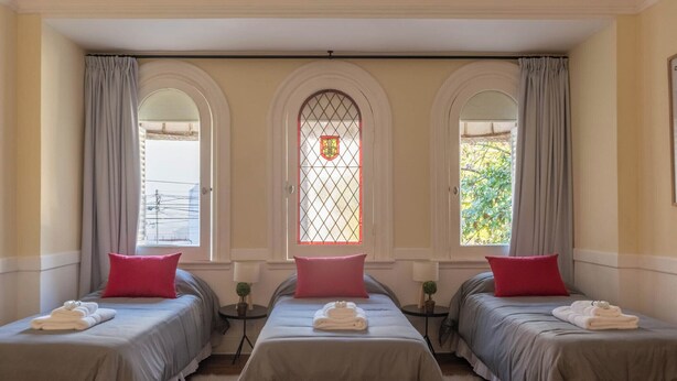 A hotel room with two beds, each with a grey bedspread and a red pillow. There are two small tables with lamps, and a window with a decorative glass panel. - MOT Mansion Paz (Buenos Aires)