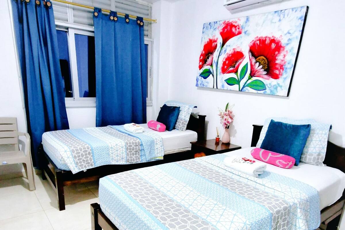 A hotel room with two beds, a window with blue curtains, a painting of red flowers, and a white air conditioning unit.