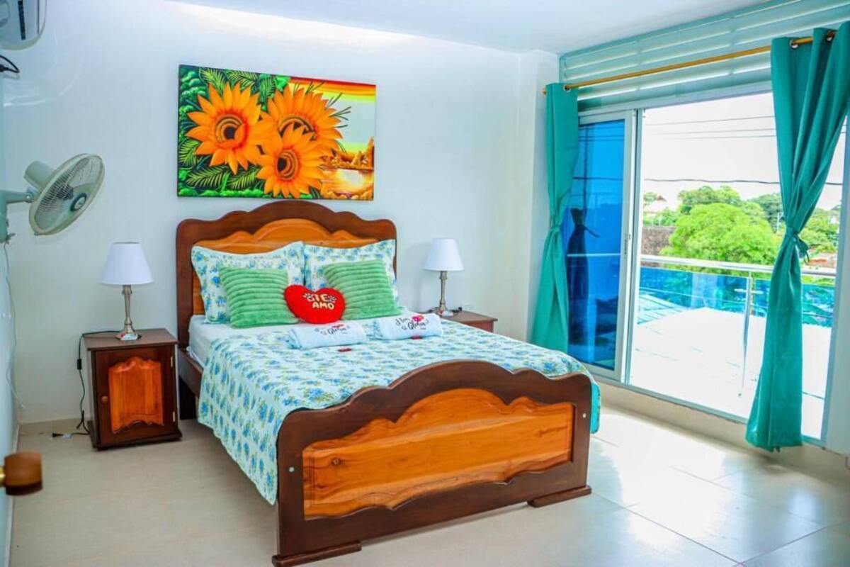 A bedroom with a wooden bed, two bedside tables with lamps, a large window with curtains, and a painting of sunflowers.