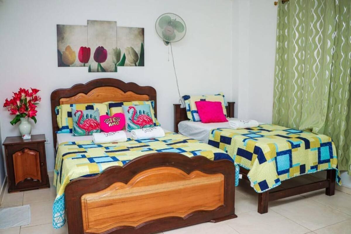 A bedroom with two beds, each with a wooden headboard and colorful bedding. There is a wall-mounted fan and a painting of tulips above the beds.