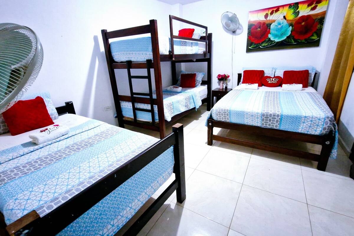 A room with two wooden bunk beds, a single bed, a standing fan, and a floral painting on the wall.