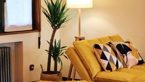 Una sala de estar con un sofá amarillo, un reposapiés blanco, una planta en maceta y una lámpara de pie.