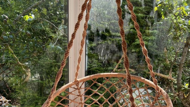 Una silla de ratán trenzado con un cojín blanco, colgada con cuerdas, ubicada junto a una ventana con vistas a un jardín exuberante.